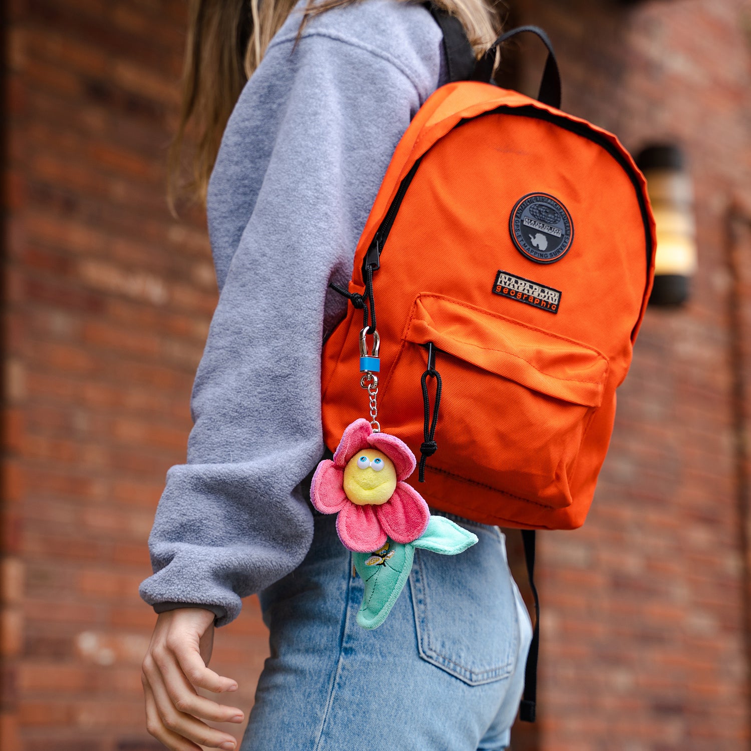 Young woman walking near a brick building wearing a sweatshirt and jeans and carrying a backpack with The Keeper Funny Friends Babee Flower AirTag holder